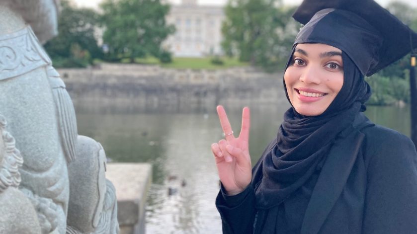 A scholar dressed in graduation garments holds up a peace sign with her fingers whilst smiling at the camera. There is a lake behind her.