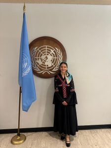 Female wearing a long black dress standing in front of a United Nations flag and sign.